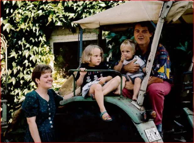 Familie mit zwei Kindern auf grünem Traktor und Frau steht daneben im Garten mit Efeu.