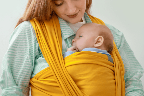 Eine Mutter trägt ihr Baby in einem gelben Tragetuch und schaut liebevoll auf das Kind.