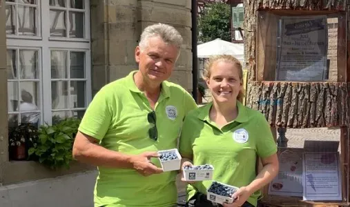 Zwei Personen in grünen Poloshirts halten Körbchen mit Heidelbeeren vor einem historischen Gebäude.
