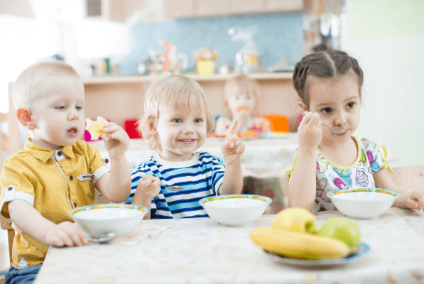 Drei kleine Kinder sitzen am Tisch und essen gemeinsam aus Schüsseln in einem hellen Raum
