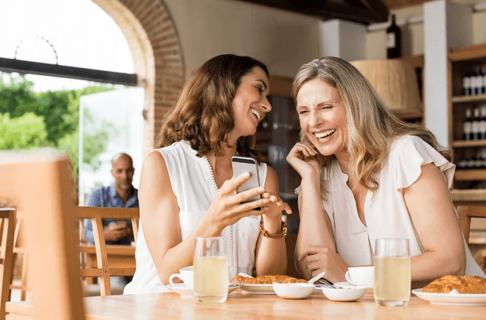 Zwei Frauen lachen beim Ansehen eines Handys in einem Café.