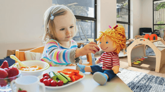 Kleines Mädchen füttert Puppe am Tisch mit buntem Gemüseteller.