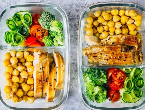 Zwei Glasbehälter mit Hähnchen, Kichererbsen, Gurken, Brokkoli und Tomaten als gesunde Mahlzeit