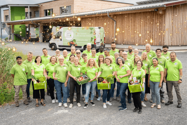Gruppenfoto von Mitarbeitern in grünen Shirts vor einem Versandlager mit Kisten und Lieferwagen