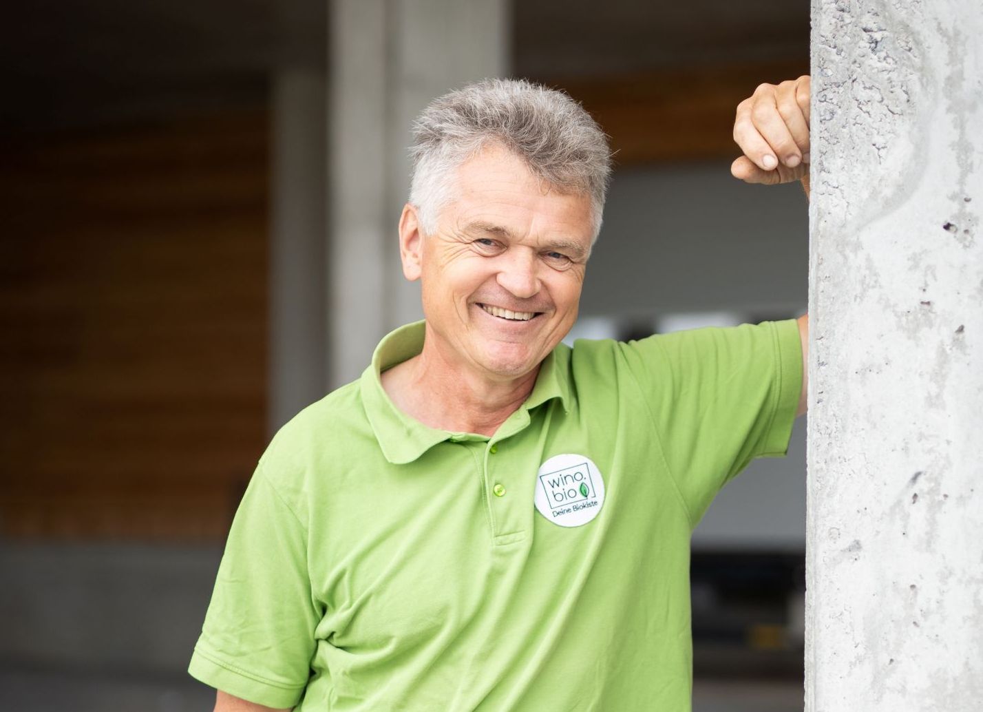 Lächelnder älterer Mann lehnt an grauer Betonwand, trägt grünes Poloshirt mit Firmenlogo