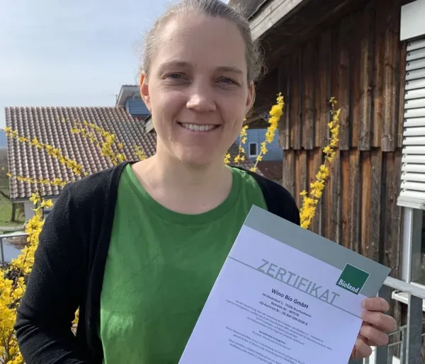 Frau hält ein Bioland Zertifikat vor einem Haus mit blühendem Busch im Hintergrund