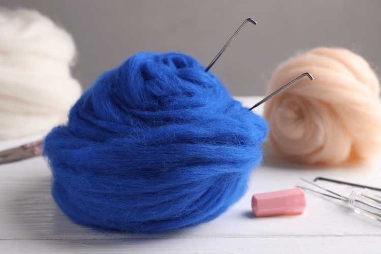 Blaue Wolle mit Nadeln zum Filzen, im Hintergrund weiße und beige Wolle und Filznadeln