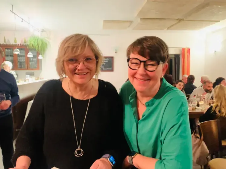 Zwei lächelnde Frauen mit Brille in einem gemütlichen Restaurant mit Gästen im Hintergrund