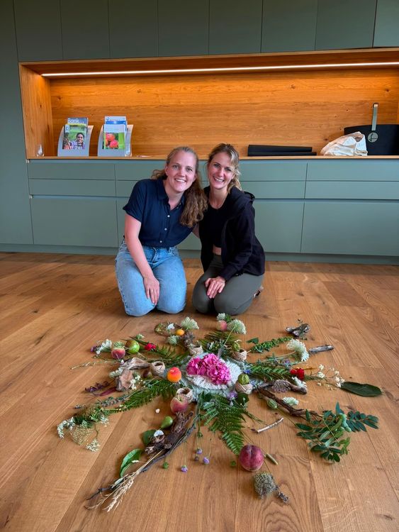 Zwei lächelnde Frauen knien vor einem kreisförmigen Kunstwerk aus Blumen und Pflanzen auf Holzboden.