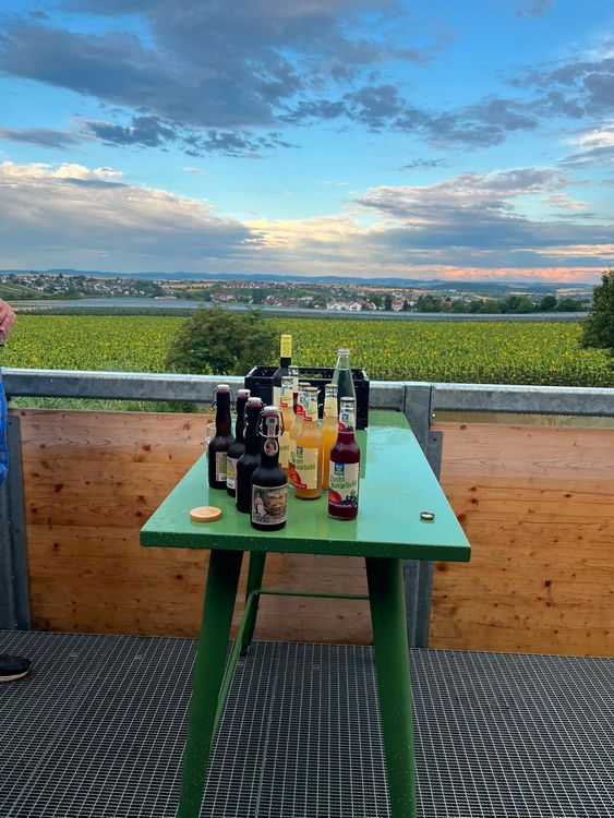 Tisch mit verschiedenen Getränkeflaschen vor Sonnenblumenfeld und Dorf unter bewölktem Himmel
