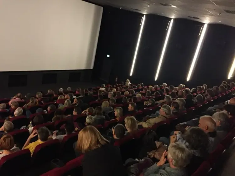 Publikum sitzt in einem dunklen Kinosaal und schaut auf eine große, leere Leinwand