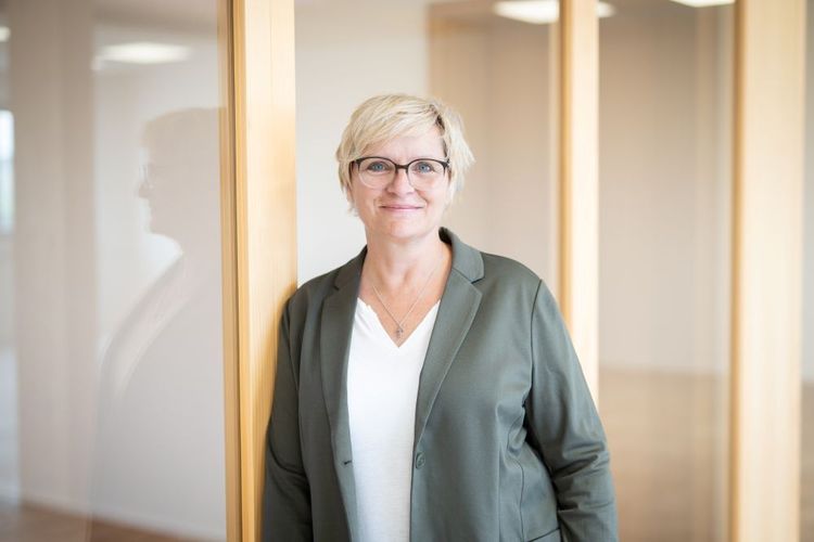 Frau mit Brille und kurzem blonden Haar lehnt an einer Glastür im Büro und lächelt freundlich.