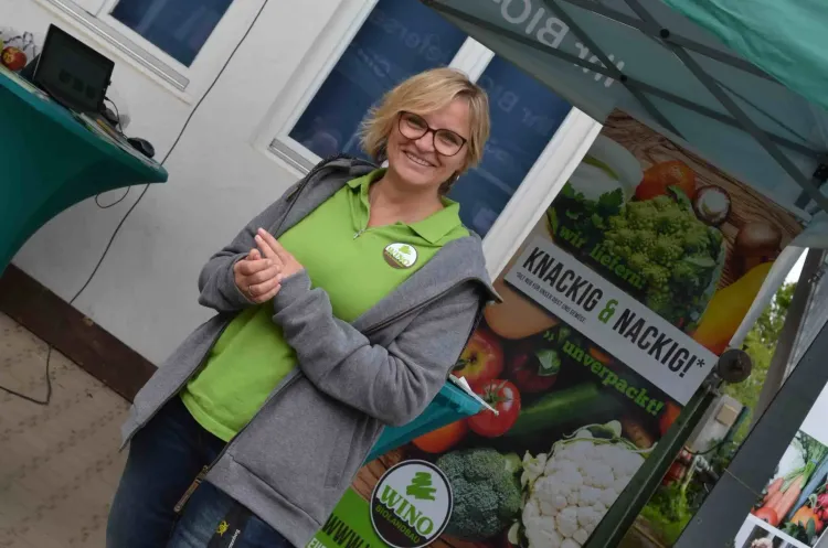 Frau in grüner Polohemd und grauer Jacke lächelnd vor Bioladen-Stand mit frischem Gemüse und Obst