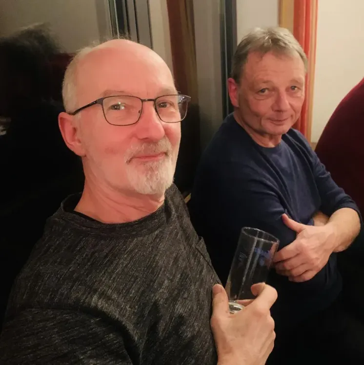 Zwei Männer, ein älterer mit Brille hält ein Glas, beide sitzen nebeneinander und lächeln freundlich
