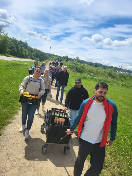 Gruppe von Menschen wandert auf einem Weg, zwei tragen Getränke und Früchte, sonniger Tag im Grünen