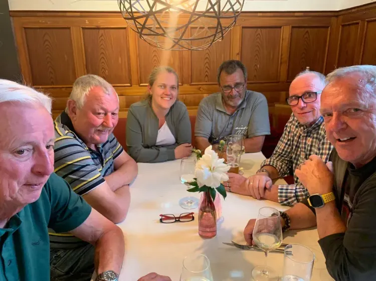Sechs Personen sitzen an einem Tisch mit weißen Blumen und trinken Wein in einem gemütlichen Raum mit Holzvertäfelung