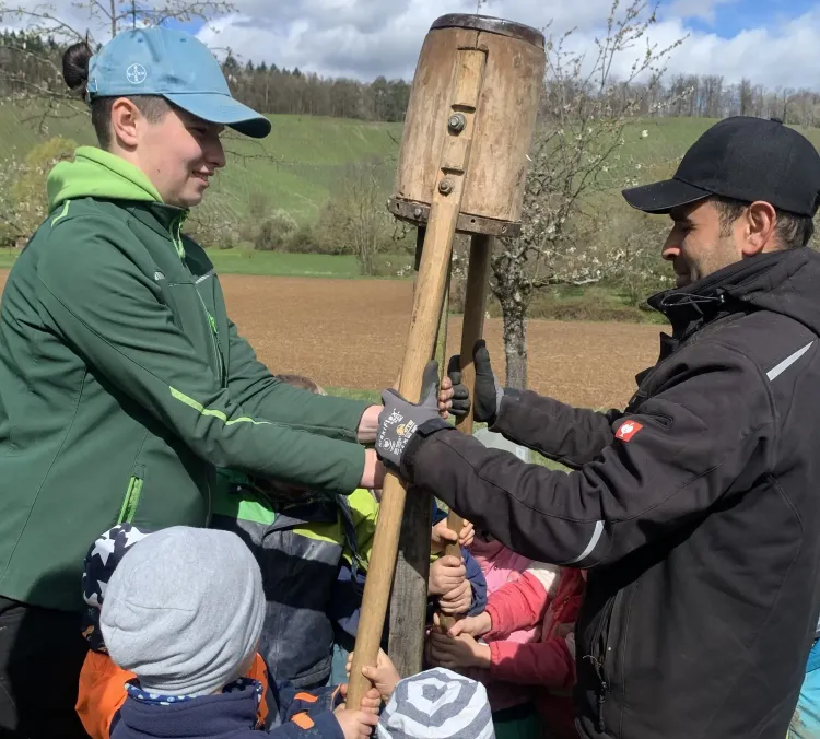 Zwei Erwachsene und mehrere Kinder halten gemeinsam einen großen Holzstampfer im Freien.