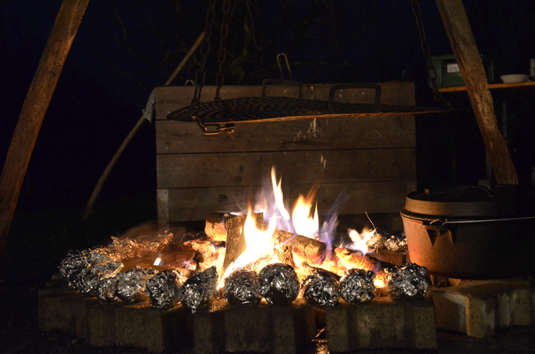 Lagerfeuer mit glühenden Kohlen und Alufolienkartoffeln, darüber ein Grillrost und ein Dutch Oven.