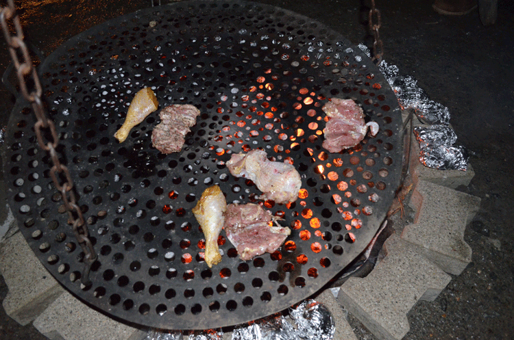 Fleischstücke und Hähnchenteile auf einem runden Grillrost über Glut mit Folienkartoffeln im Hintergrund.