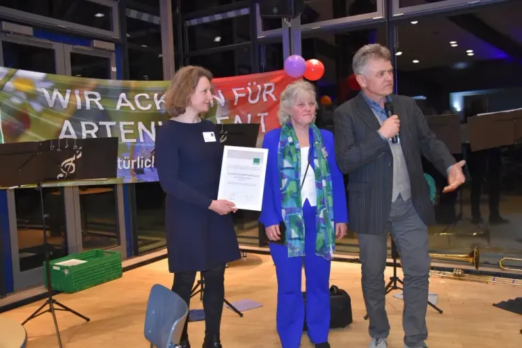 Drei Personen bei Auszeichnung vor Banner für Artenschutz in Veranstaltungsraum