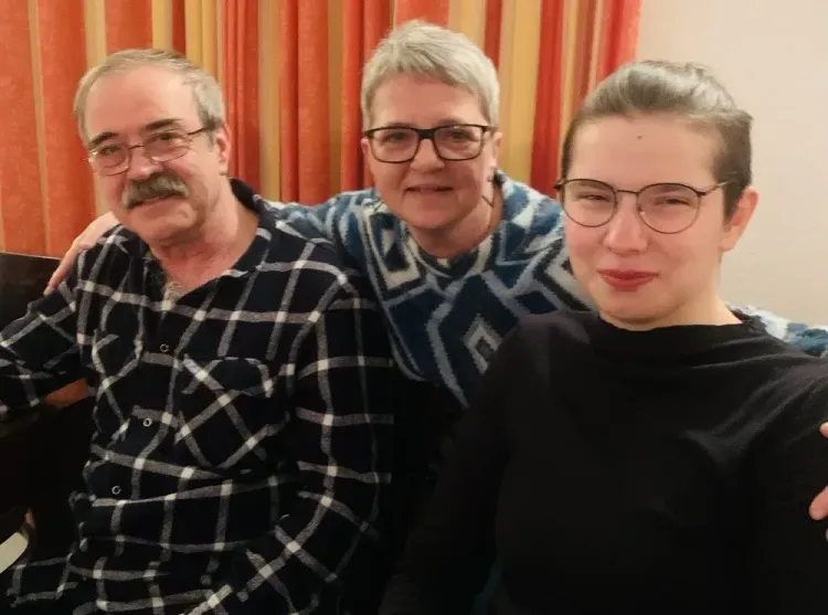 Drei Personen mit Brillen sitzen eng zusammen und lächeln vor orangenen Vorhängen