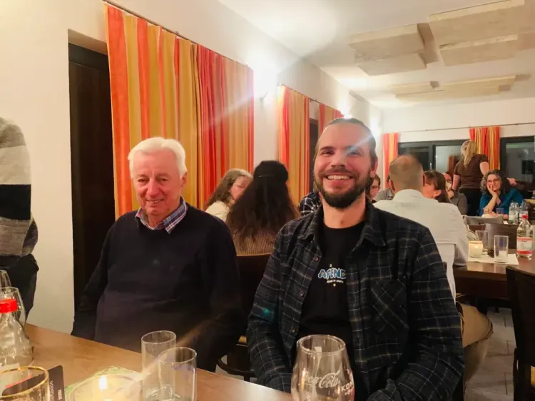 Zwei Männer unterschiedlichen Alters lachen an einem gedeckten Tisch in einem Restaurant mit gelb-roten Vorhängen.