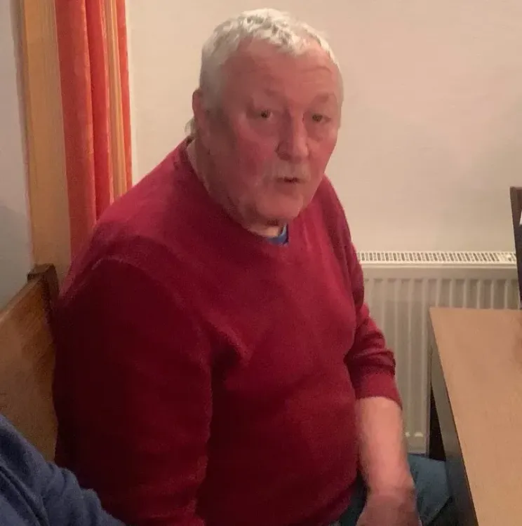 Älterer Mann mit rotem Pullover sitzt an einem Holztisch in einem Innenraum