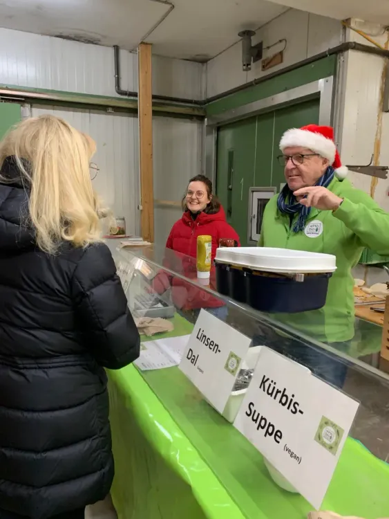 Drei Personen am Essensstand mit Schildern Linsen-Dal und Kürbissuppe, Mann mit Weihnachtsmütze