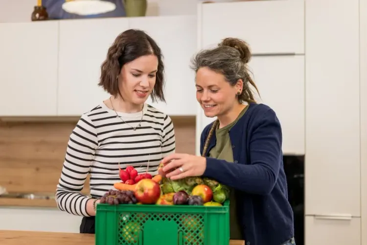 Zwei Frauen in einer Küche betrachten gemeinsam frisches Obst und Gemüse in einer grünen Kiste.