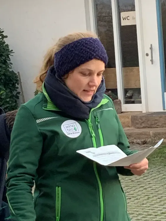 Frau in grüner Jacke und Stirnband liest ein Blatt Papier vor einem Gebäude mit WC-Schild