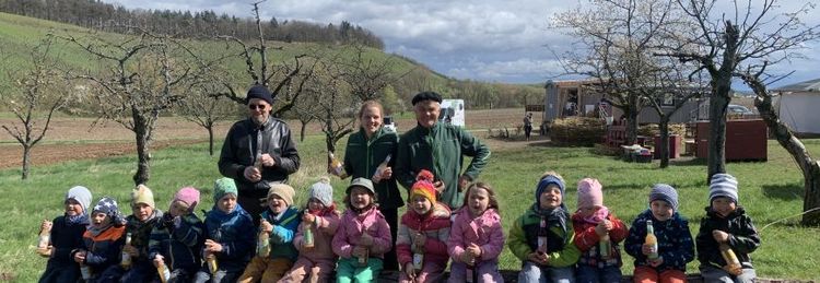 Kinder und zwei Erwachsene sitzen draußen auf einer Wiese und trinken Saft, Bäume und Hügel im Hintergrund