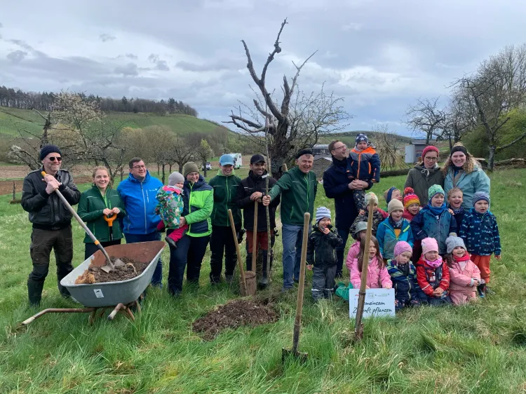 Gruppenfoto von Erwachsenen und Kindern beim Bäumepflanzen auf einer Wiese mit Werkzeug und Schubkarre