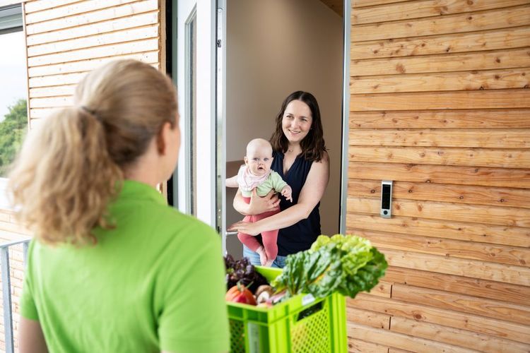 Eine Frau hält ein Baby in der Tür, eine andere Frau liefert frisches Gemüse in einer grünen Kiste