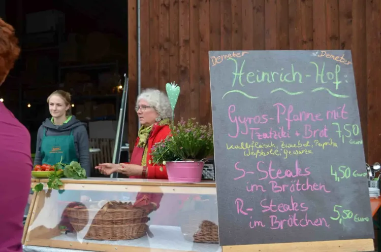 Zwei Frauen an einem Marktstand mit einer Speisekarte von Heinrich-Hof und frischem Gemüse.