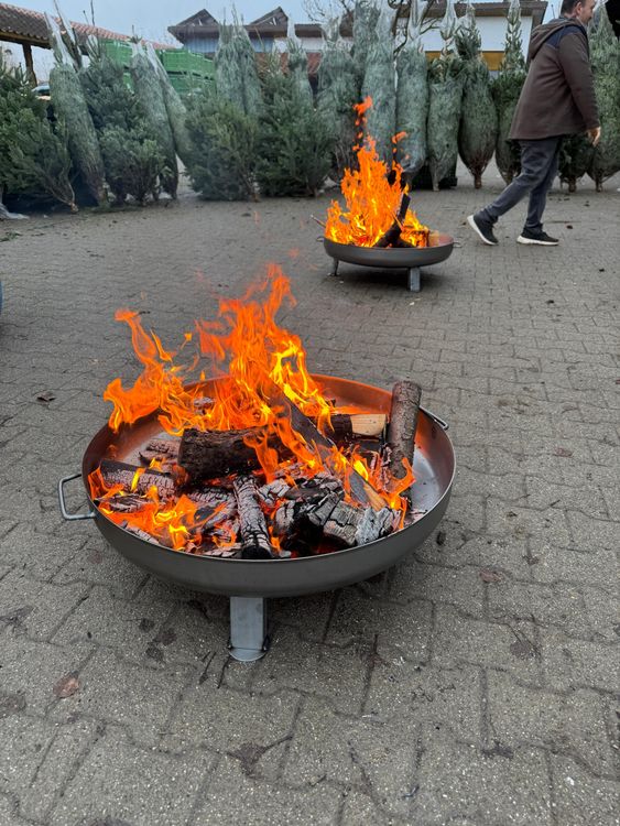 Flammen in zwei Feuerschalen vor aufgereihten Weihnachtsbäumen und einem gehenden Mann.