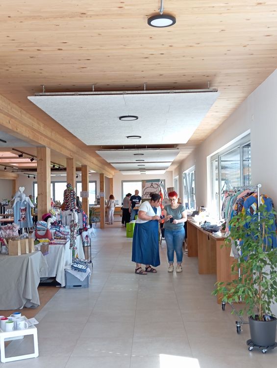 Innenansicht eines Marktplatzes mit Holzdecke, Verkaufsständen und Menschen beim Einkauf und Gespräch