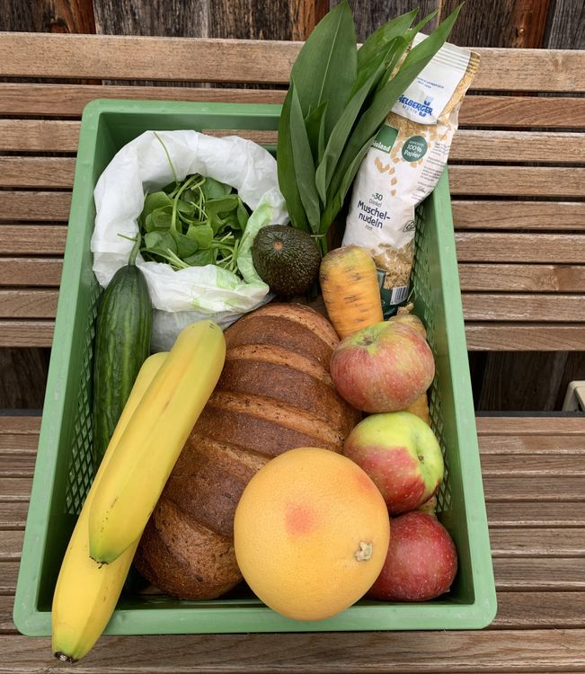 Grüner Korb mit Brot, Bananen, Gurke, Äpfeln, Grapefruit, Avocado, Karotte, Wildkräutern und Dinkelnudeln