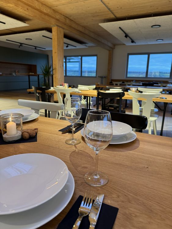 Modernes Restaurant mit gedecktem Holztisch, Kerze und Weingläsern vor großen Fenstern und hellen Stühlen