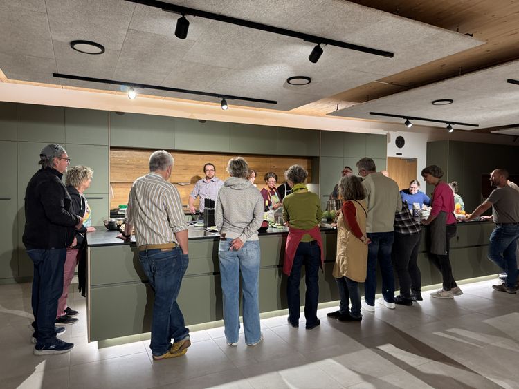 Menschen stehen an einer Kücheninsel, während eine Person in der Mitte etwas erklärt oder kocht.