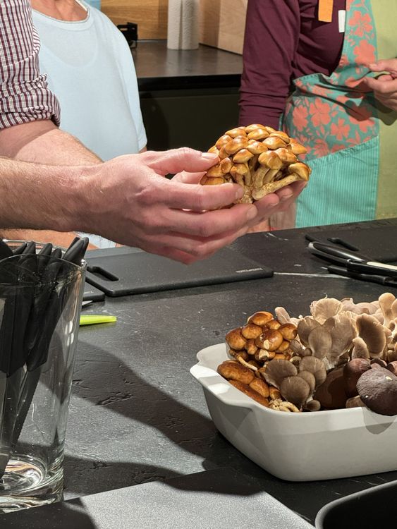 Zwei Personen in der Küche, die einen großen Pilz in den Händen halten, mit weiteren Pilzen in einer Schale auf der Arbeitsfläche