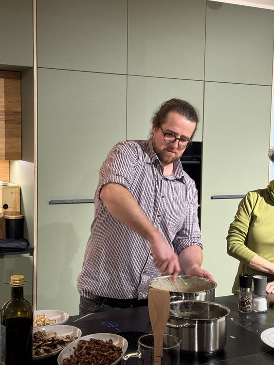 Mann mit Brille rührt in einem Topf in einer modernen Küche, Zutaten und Gewürze auf der Arbeitsplatte