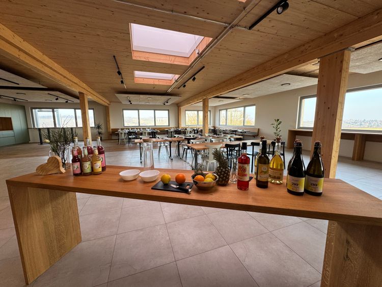 Großer heller Veranstaltungsraum mit Holztischen und Stühlen, Buffet mit Getränken und frischem Obst