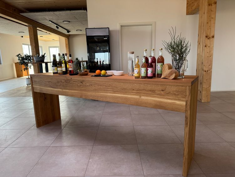 Moderner Holztisch mit verschiedenen Getränkeflaschen, Obst und Dekoration in hellem Raum mit Holzstützen