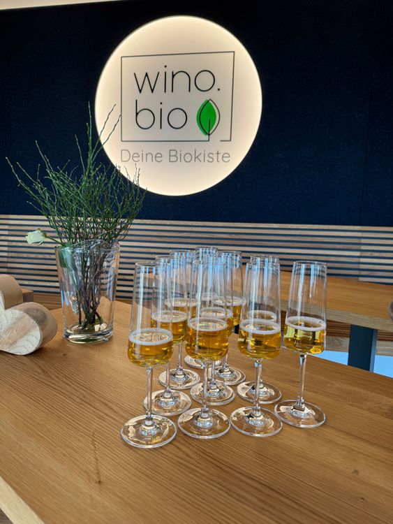 Gläser mit Getränken auf einem Holztisch vor beleuchtetem wino.bio Logo mit grünem Blatt.