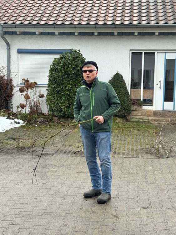 Mann mit grüner Jacke, Jeans und Sonnenbrille hält einen Ast vor einem Haus mit weißer Fassade