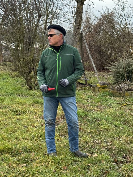 Älterer Mann im grünen Jacke und Jeans steht draußen auf Grasfeld mit Handschuhen und Sonnenbrille