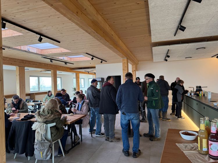 Menschen in einem modernen Café mit Holzdecke und großen Fenstern, einige sitzen, andere stehen und unterhalten sich
