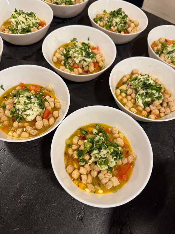 Mehrere weiße Schalen mit einer Suppe aus Gemüse, Kräutern und kleinen Croutons auf dunklem Untergrund.