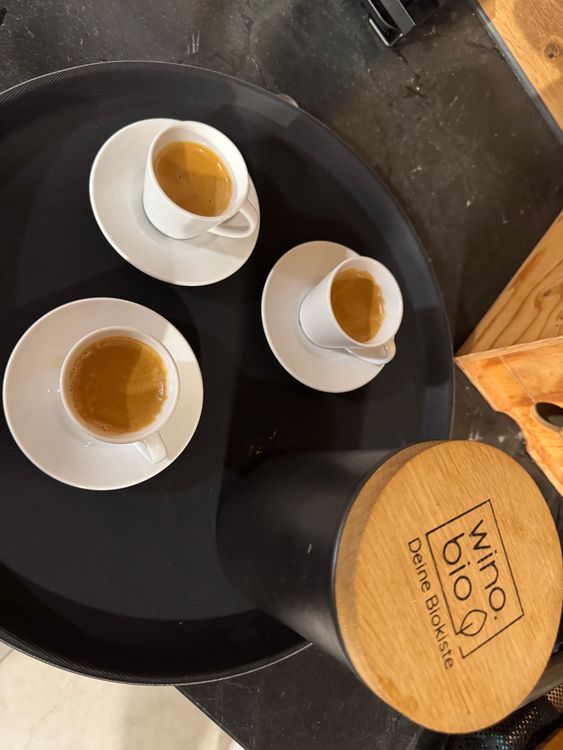 Drei Espressotassen mit Kaffee auf einem schwarzen runden Tablett neben einem Holzkübel mit der Aufschrift wino bio