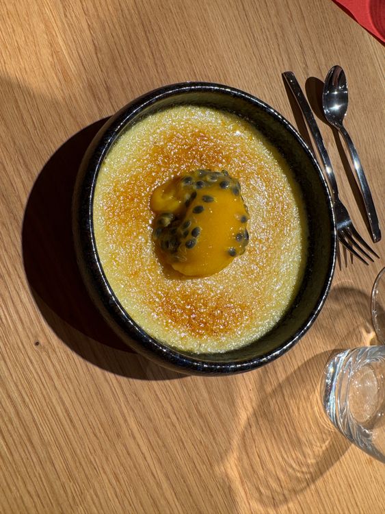 Creme Brulee Dessert mit Passionsfruchtsoße in schwarzer Schale auf Holztisch mit Besteck und Glas